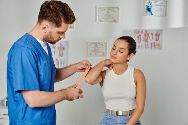 Tıbbi kostümlü yakışıklı doktor kadın hastasının vücuduna kinesiyolojik bantlar yapıştırıyor.