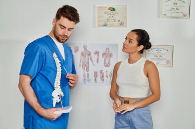 Mavi tıbbi kostümlü yakışıklı doktor, omurga modelini genç bayan hastasına gösteriyor, sağlık hizmetleri.