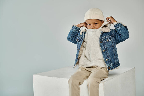 cute african american boy in winter attire and beanie sitting on concrete cube and adjusting hood