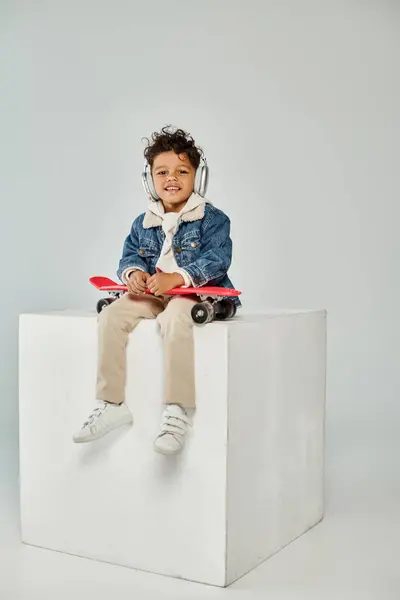 stock image cute african american boy in winter attire and headphones sitting on cube with penny board on grey