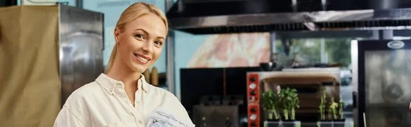 happy blonde woman working as manager looking at camera in cozy and modern cafe, horizontal banner
