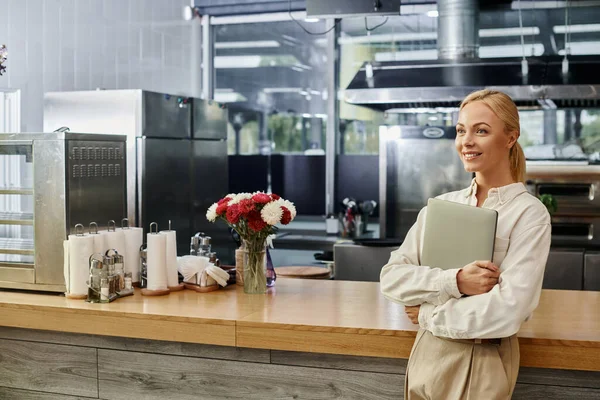 cheerful blonde cafe administrator with laptop smiling and looking away near counter, small business