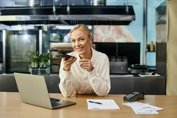 金发女人用智能手机发送语音信息 并在咖啡店的笔记本电脑旁对着摄像机微笑 — 图库照片