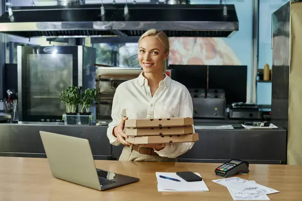 smiling blonde woman with pizza boxes near digital devices and payment terminal on counter in cafe