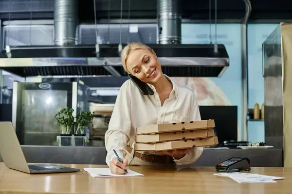 happy blonde woman with pizza boxes talking on smartphone and writing order on counter in cafe