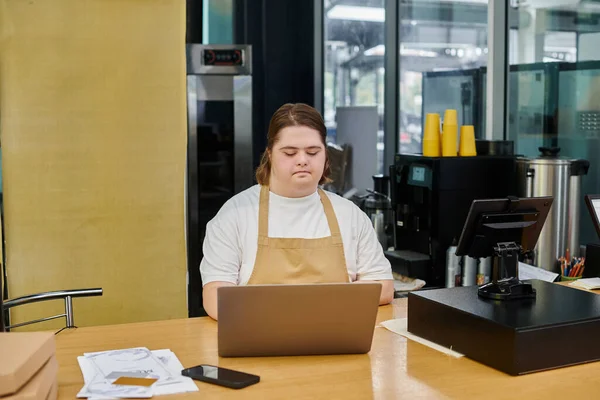 concentrated young woman with down syndrome working on laptop on counter in contemporary cafe