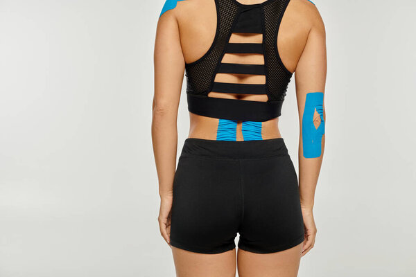 cropped view of young woman in black sport attire with kinesiological tapes on back on gray backdrop