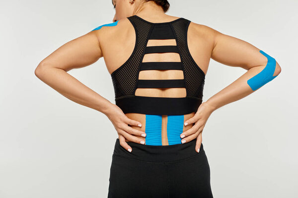 back view of young woman in black sport wear posing on gray background with kinesio tapes on body