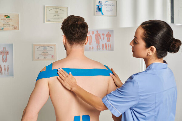 beautiful young doctor putting kinesiological tapes on back and shoulders of her patient, healthcare