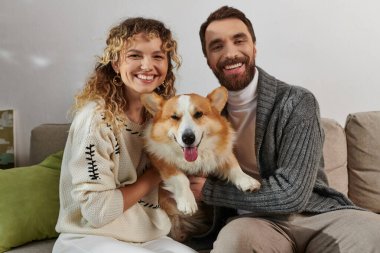 Kış kıyafetleri içinde neşeli bir çift modern apartman dairesinde gülümsüyor ve Corgi köpeğiyle oynuyor, mutlu anlar.