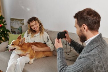 Mutlu adam gülümsüyor ve karısının Corgi Dog 'la Noel ağacının yanında fotoğrafını çekiyor.