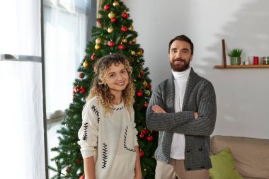 happy couple in winter clothing standing near decorated Christmas tree in modern apartment clipart