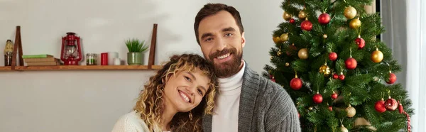 stock image joyful couple in winter clothing standing near decorated Christmas tree in modern apartment, banner