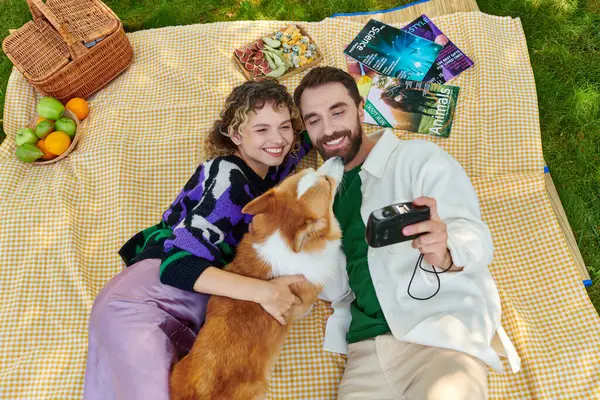 stock image top view of happy couple taking photo with cute corgi dog on camera while having picnic in park