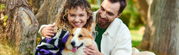happy man hugging curly woman in outfit while cuddling corgi dog in park, sitting near tree banner