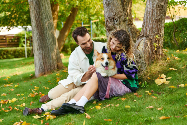 happy man and curly woman in cute outfit cuddling corgi dog and sitting near tree in autumnal park