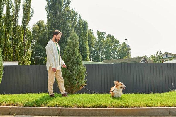 happy bearded man in cozy warm attire walking with cute corgi dog on green lawn near home