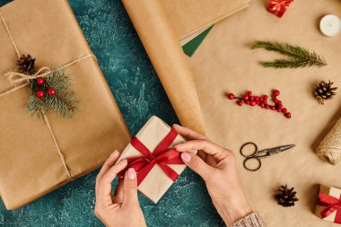 Hediye kutusuna kırmızı fiyonk bağlayan bir kadının Noel hediyesini dekorasyon malzemelerinin yanında hazırlarken görüntüsü.