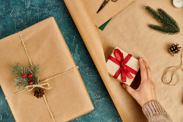 cropped view of woman with decorated gift box near craft paper and Christmas decor on blue backdrop