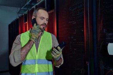 Güvenlik yeleği giymiş sakallı yakışıklı teknisyen ve eldivenli detektör ve telefonla konuşuyor.
