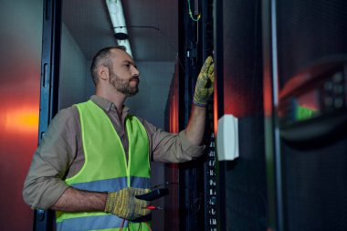 pensive specialist in safety vest and gloves using detector during inspection of data center clipart