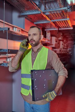 Güvenlik yeleği ve eldivenli yakışıklı dalgın bir adam çok çalışırken akıllı telefonla konuşuyor.