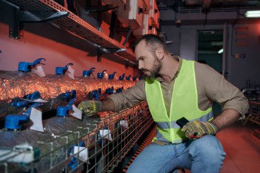 handsome concentrated specialist in safety vest and gloves squatting while working in data center clipart