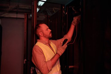 concentrated devoted technician in safety vest and gloves using detector in data storage during work clipart