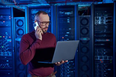 pensive data center specialist in turtleneck with glasses talking by phone and looking at his laptop clipart