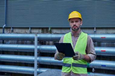 Kask ve güvenlik yeleği giymiş odaklanmış teknisyen elinde bir pano tutuyor ve kameraya, veri merkezine bakıyor.