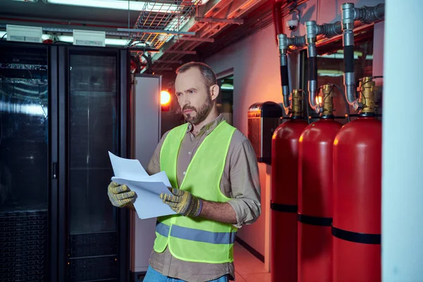 stock image good looking pensive specialist in safety clothes holding papers and looking away, data center