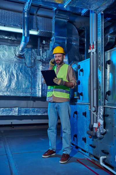 веселый преданный техник в защитной одежде и шлеме смотрит в буфер обмена и разговаривает по телефону