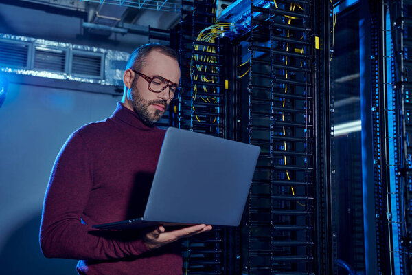 good looking pensive data center specialist in turtleneck with glasses and beard looking at laptop