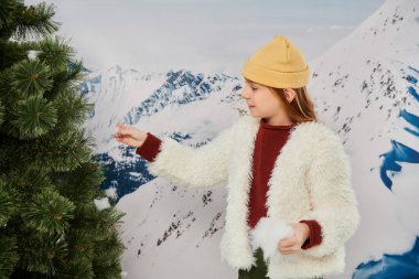 joyful pretty girl in beanie hat and warm attire putting snow on fir tree, fashion concept clipart