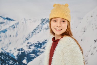 portrait of stylish little girl in warm trendy outfit smiling at camera with mountain backdrop clipart