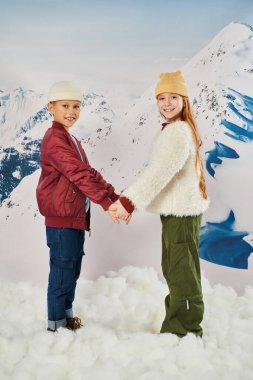 cute preteen friends in stylish winter jackets and beanie hats holding hands and smiling at camera clipart