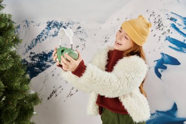 Kış kıyafetli mutlu ergen kız köknar ağacının yanında kulaklığına bakıyor, moda konseptine.