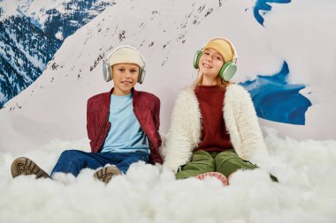 Şık kış kıyafetleri içinde kulaklıklarla karda oturan neşeli moda arkadaşları, moda kavramı.