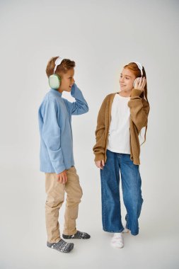jolly preadolescent friends posing with headphones on gray backdrop smiling at each other, fashion clipart