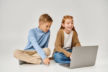 joyous preteen boy and girl sitting on floor and looking happily at laptop, fashion concept clipart