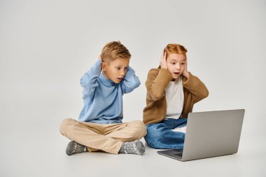 two scared trendy little children looking at laptop and closing ears with hands, fashion concept clipart