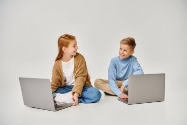 joyous preadolescent friends sitting on floor with crossed legs with laptops smiling at each other clipart