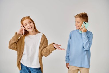 Keyifli, modaya uygun giyinmiş, gri arka planda telefonla konuşan ergenlik öncesi çocuklar.