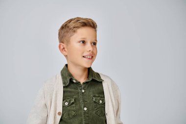 Sıcak bir hırka giymiş neşeli küçük bir çocuk. Gülümsüyor ve gri arka plana bakıyor. Moda konsepti.
