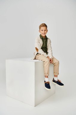 Moda hırkalı, beyaz küpün üzerinde oturan ve kameraya gülümseyen küçük çocuğun dikey görüntüsü.