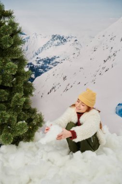 cheerful red haired little girl playing with snow next to fir tree with mountain on backdrop clipart