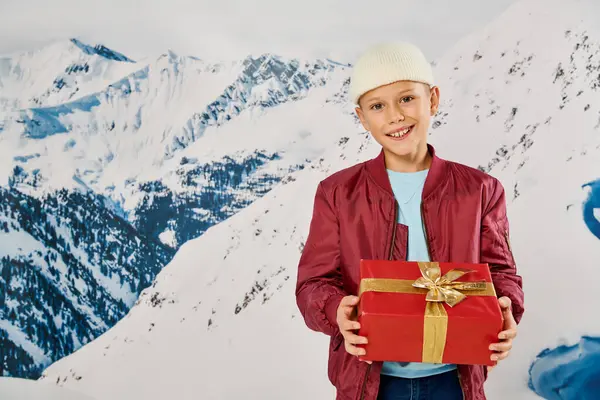 stock image joyful preadolescent boy in stylish winter attire holding present and smiling at camera, fashion