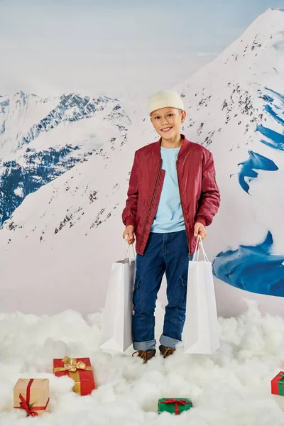 stock image cheerful preteen boy in red stylish jacket holding present bags and smiling at camera, fashion