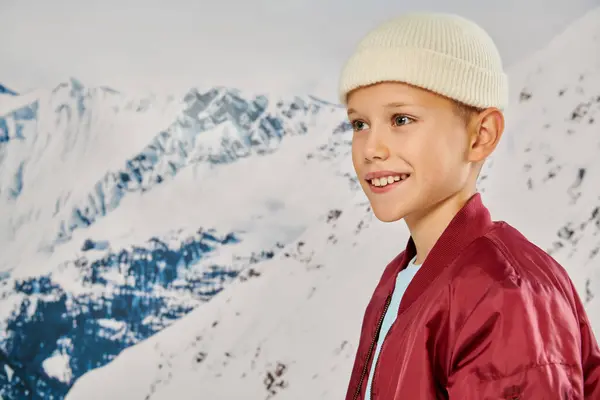 stock image portrait of jolly preteen boy in beanie hat looking away with snowy mountain on backdrop, fashion