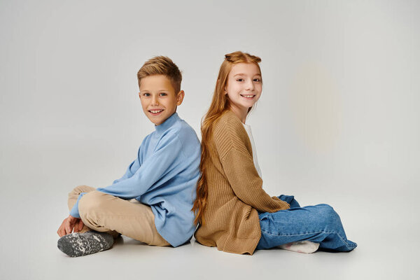 joyful preteen children sitting on floor back to back with crossed legs smiling at camera, fashion